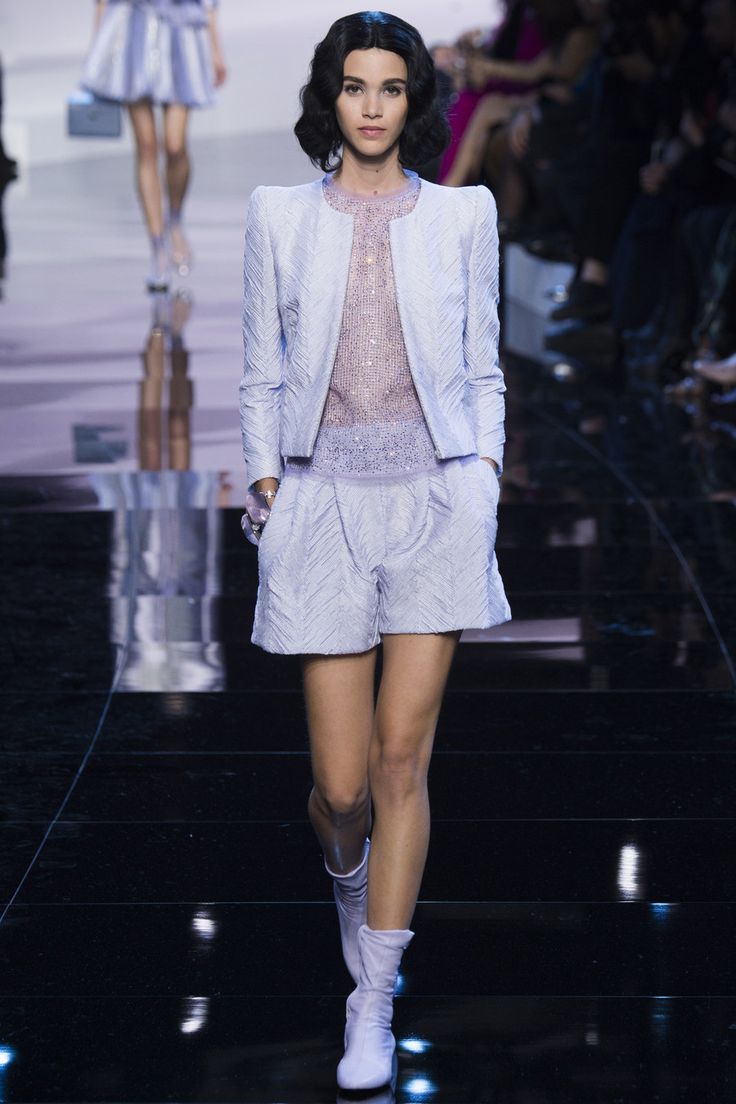 a female model walks down a runway wearing a white jacket and matching shorts, with white boots. the model is posed in a fashionable way, with her hair styled, and her accessories adding to the overall look. the runway is black and shiny, highlighting the outfit's style and glamour.  the model looks confident and elegant.