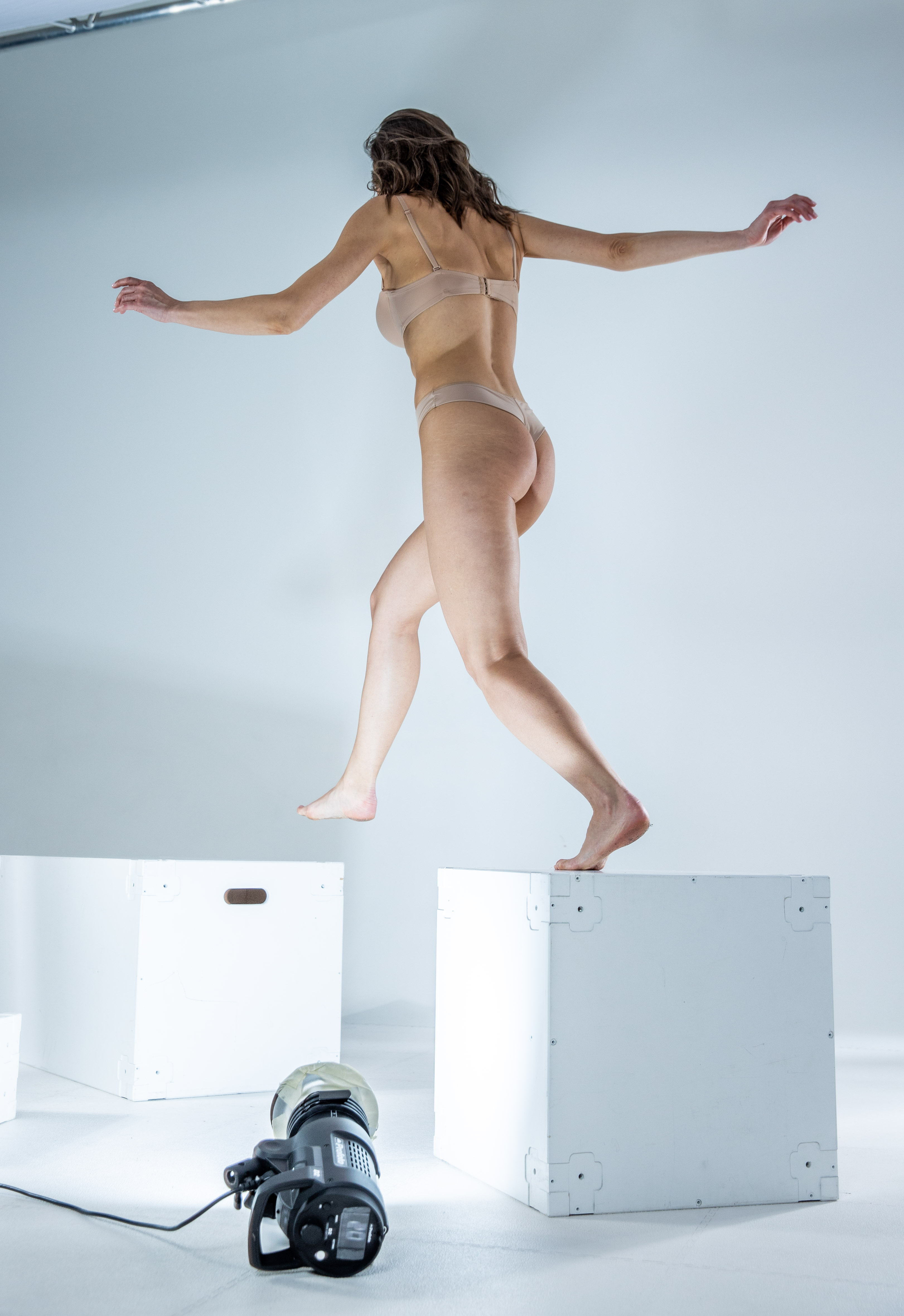 a woman wearing underwear is captured midjump between white boxes in a studio setting, with lighting equipment visible in the foreground.