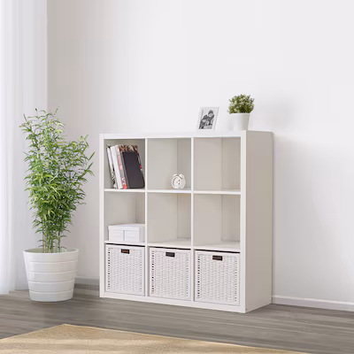 an indoor shot shows a white cube shelving unit with six cubbies. the unit is placed on a gray wood floor, next to a beige rug. in the bottom row, three woven baskets fill three of the cubbies. the top left cubby contains books and a small white box is in the middle left cubby. on top of the unit are a small framed photo and a potted plant. a potted plant sits on the floor, to the left of the shelf. a white curtain hangs in the background.
