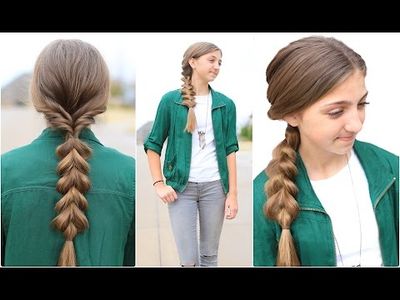a young girl is demonstrating a hairstyle. the girl is wearing a green jacket over a white tshirt and a pair of gray jeans. she is standing on a sidewalk with green grass on either side. the lighting is natural daylight. the perspective of the image is eye level.