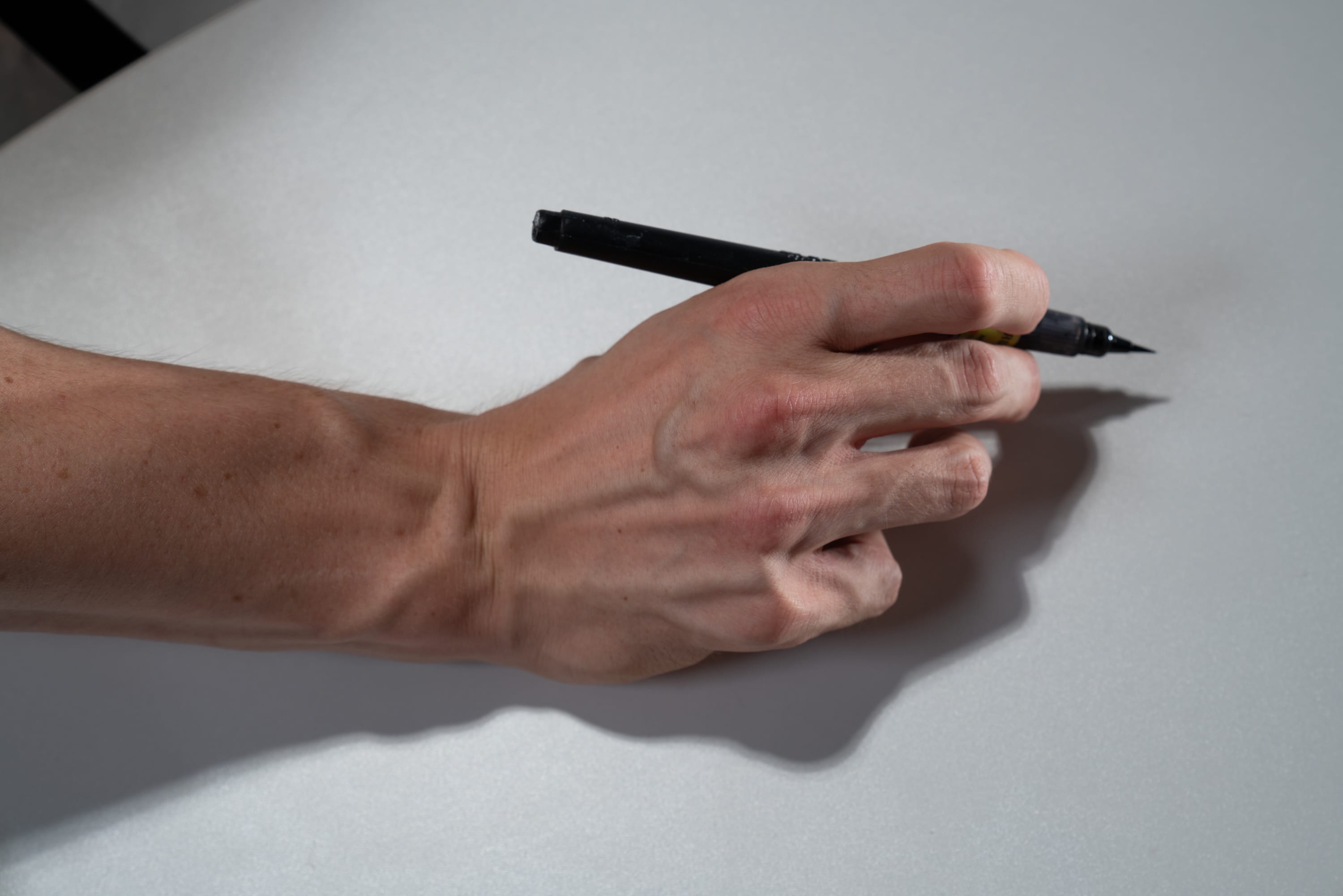 a caucasian man's hand is shown holding a black marker, poised to write on a white table. the image is a closeup, showing the veins and details of the hand. the lighting is natural, with a shadow cast by the hand.