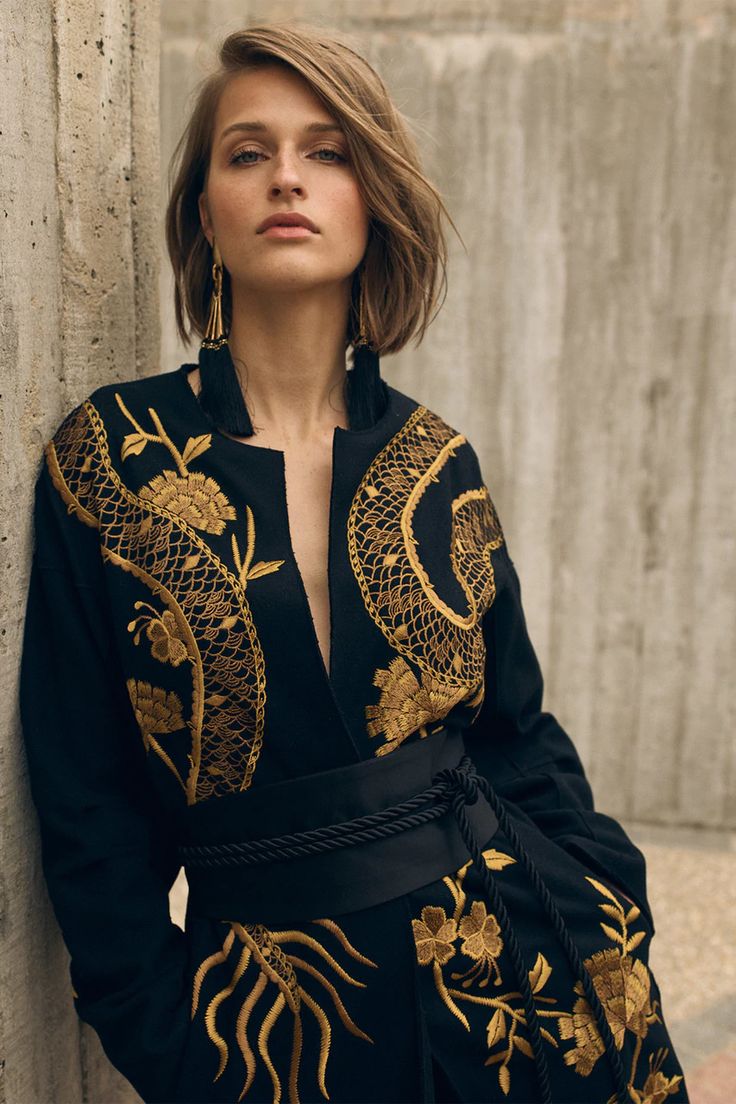 loseup view of a fashion model wearing a stylish black dress adorned with intricate gold embroidery. the dress features floral and possibly dragonlike patterns. the model has shoulderlength brown hair and is wearing gold tassel earrings. she is posing against a textured concrete wall. the overall aesthetic is elegant and sophisticated, suggestive of a fall or autumn fashion collection. the image highlights the detail of the embroidery and the overall look of the dress.