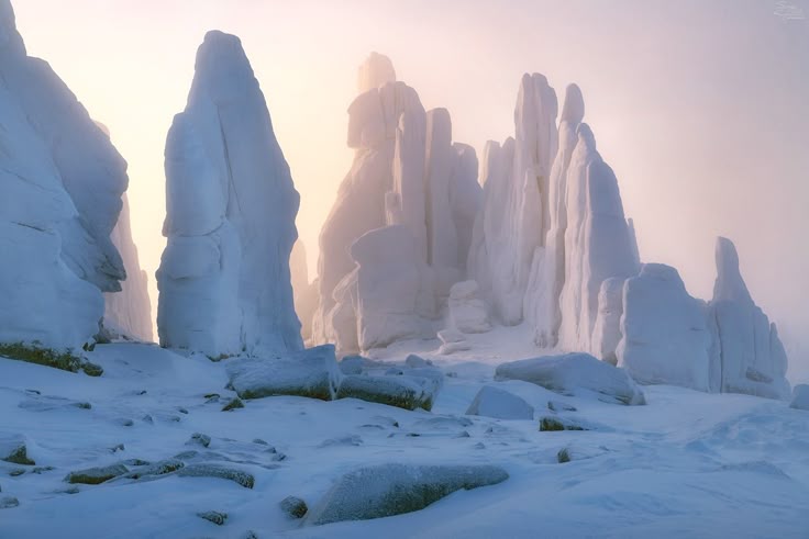 the image shows a snowy landscape with large rock formations covered in snow and ice. the rocks are tall and pillarlike, and they create a dramatic scene. the ground is also covered in snow, and a fog or mist hangs in the air, softening the light and creating a sense of mystery. the overall feeling is one of coldness and isolation.
