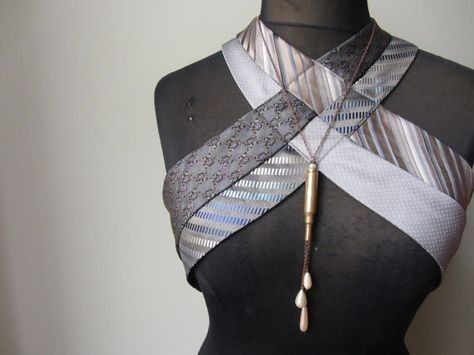 a mannequin is wearing a neckpiece made of repurposed neckties. the ties are arranged in a crisscross fashion around the mannequin's upper torso. a necklace with a bullet pendant and dangling pearls adorns the neckpiece. the ties are in shades of grey and silver with various patterns, including stripes and geometric designs.