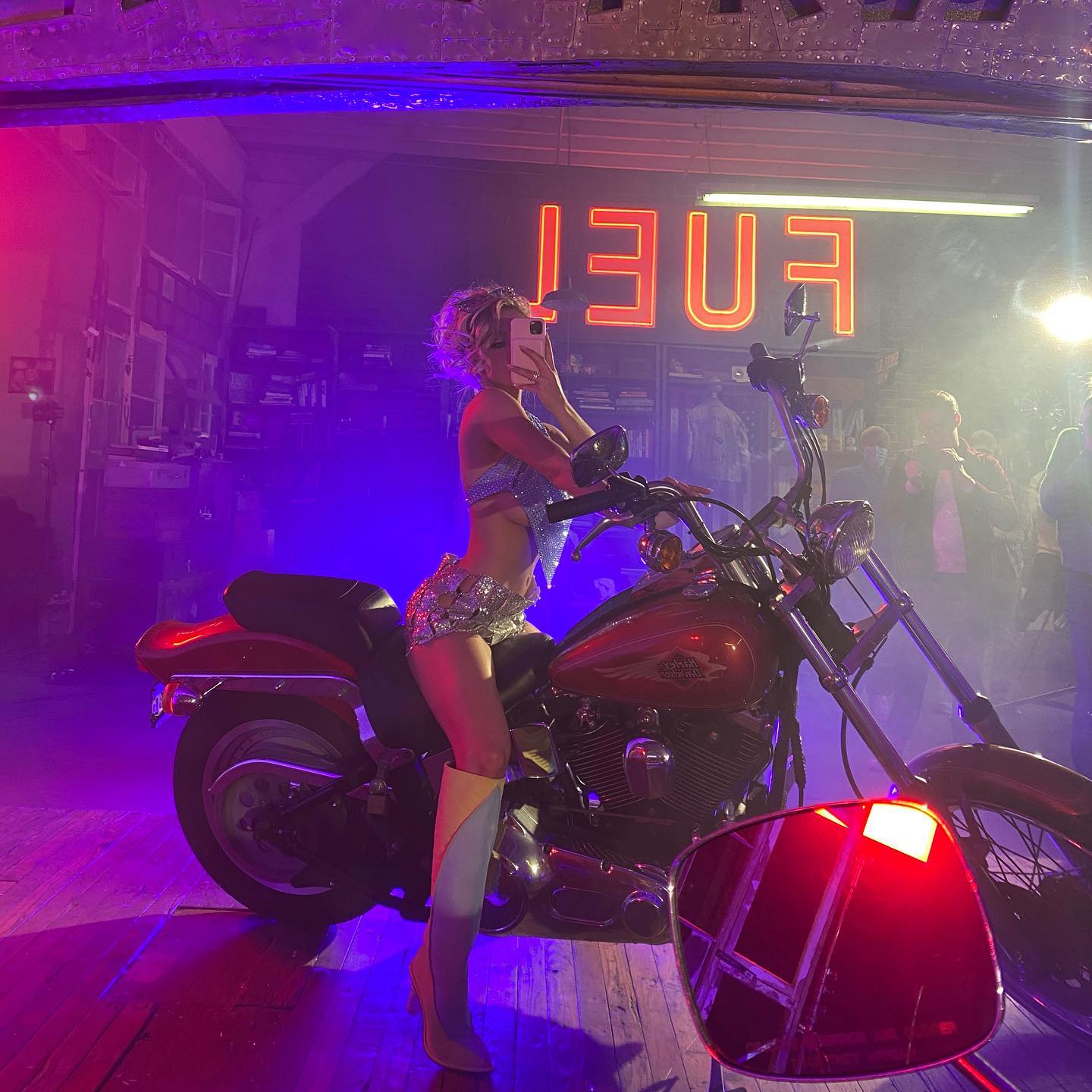 the image shows a woman in a sparkly, light blue and silver twopiece costume and high platform boots posing on a red harley davidson motorcycle.  the setting appears to be a dimly lit studio or stage, bathed in purple and pink neon lighting. a neon "fuel" sign is visible in the background. the atmosphere suggests a behindthescenes shot from a photoshoot or film production.  other people are blurred in the background, implying a crew or onlookers. the overall aesthetic is glamorous and retro.