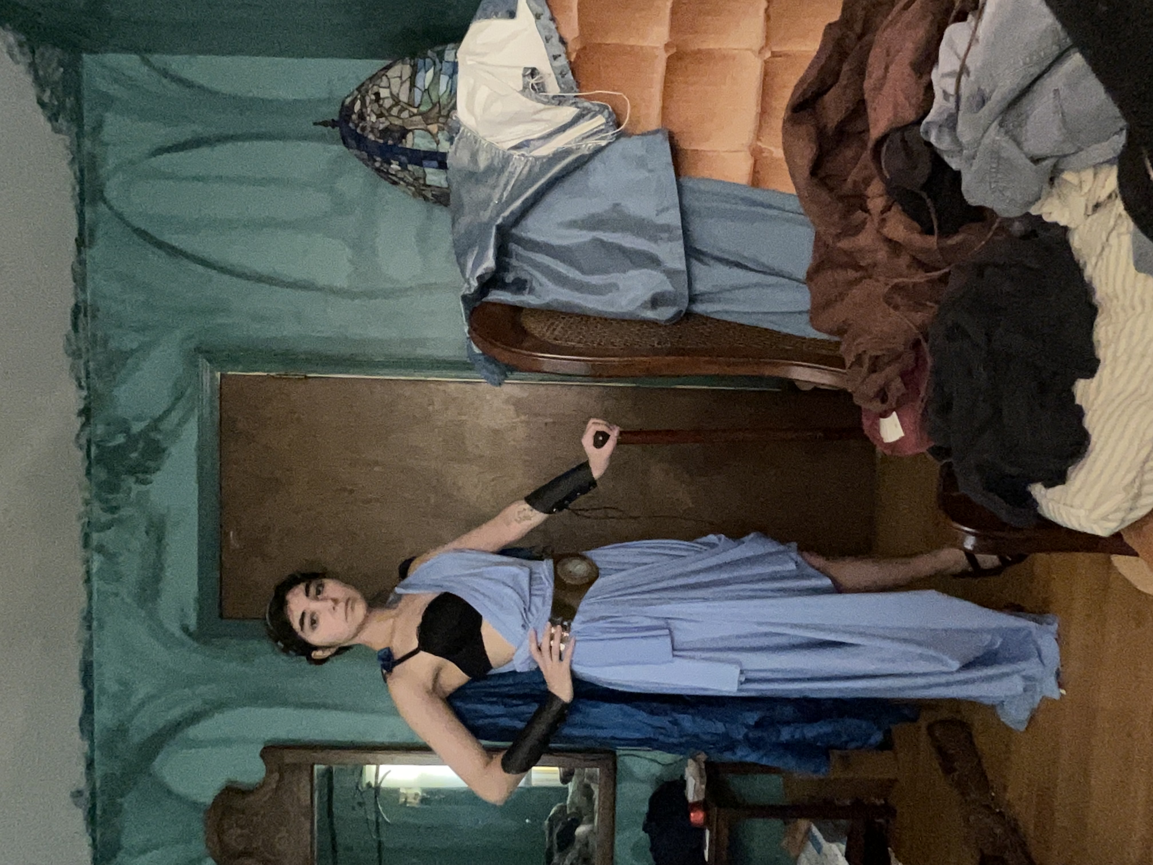 a fullbody shot of a woman dressed in a light blue grecianstyle costume. she's standing in a messy bedroom with teal walls and vintage furniture. the woman holds a cane in one hand, and her costume features a draped dress, arm bands, and a headband. the background includes a cluttered chair and bed, showcasing a generally messy and livedin space. the overall feel is a blend of historical costume and contemporary casual setting.