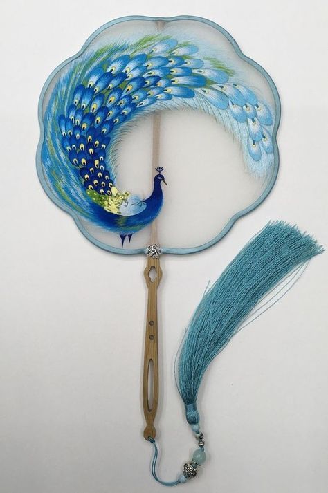 a beautiful hand embroidered silk fan with a peacock design. the fan is blue and white with a wooden handle and a blue silk tassel.