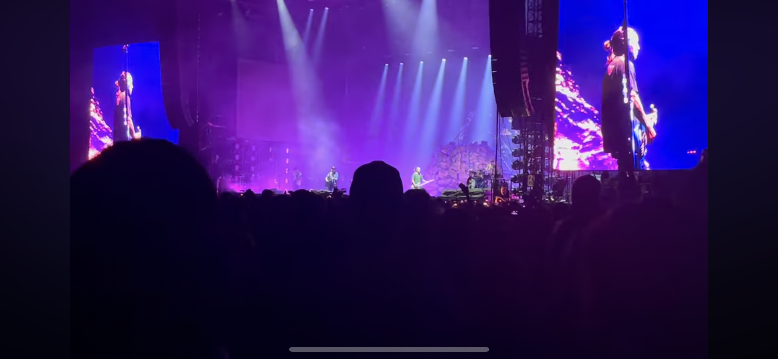 a concert with a band on stage performing for a crowd of fans, the lights are on the band and the stage, the back of the crowd is facing the camera. the band is in silhouette and there are two large screens on either side of the stage.  the image is dark with purple lighting and the silhouette of a person is in the foreground.
