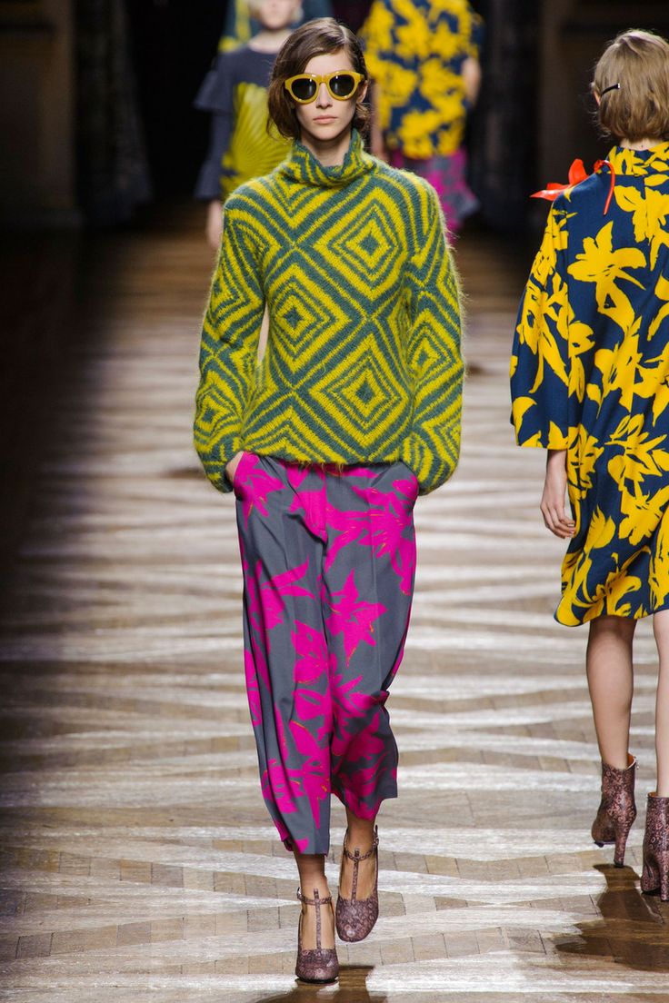 a runway model walks down the catwalk wearing a striking outfit.  the outfit consists of a geometric patterned yellow and green sweater, grey and hot pink floral print pants, and strappy heeled shoes.  she is wearing yellow sunglasses. another model, wearing a bold floral dress, walks behind her. the overall aesthetic is one of vibrant color and bold patterns. the runway floor is a polished wood.