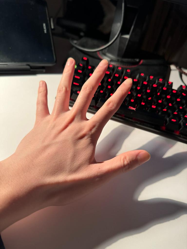 a pale hand is outstretched on a white desk in front of a black computer keyboard with red backlighting. the hand is positioned palm up, with fingers slightly spread, showcasing prominent blue veins against the fair skin. the image is taken from a firstperson perspective, suggesting the owner of the hand is sitting at the desk. the natural lighting creates shadows on the desk, highlighting the hand's contours and the keyboard's keys.