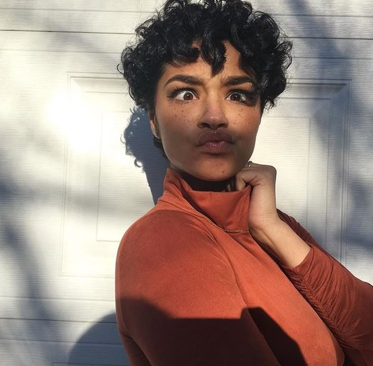 a woman with curly black hair and freckles is making a funny face with crossed eyes and puckered lips. she is wearing an orange turtleneck and standing in sunlight with a shadow behind her. she is in front of a white door.