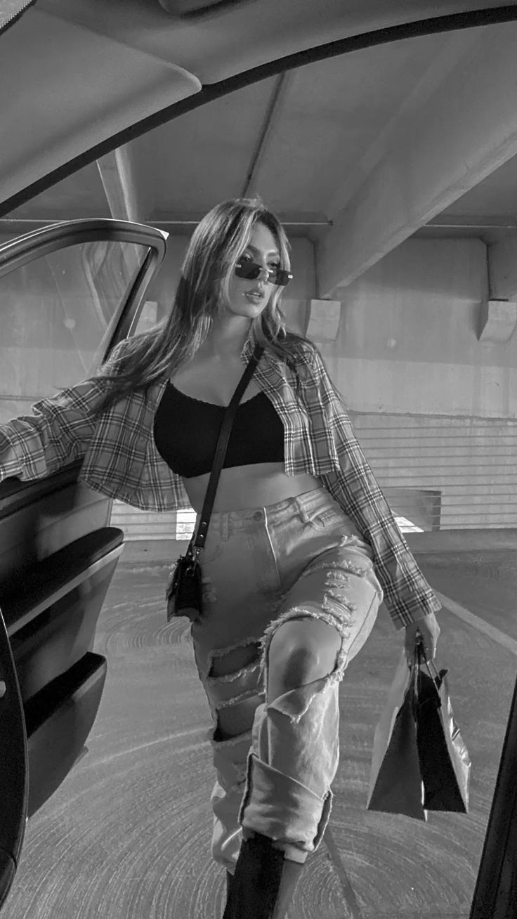 a black and white photo of a stylish young woman leaning against a car in a parking garage. she's wearing a plaid shirt, ripped jeans, sunglasses, and a handbag. the overall aesthetic is cool and trendy.