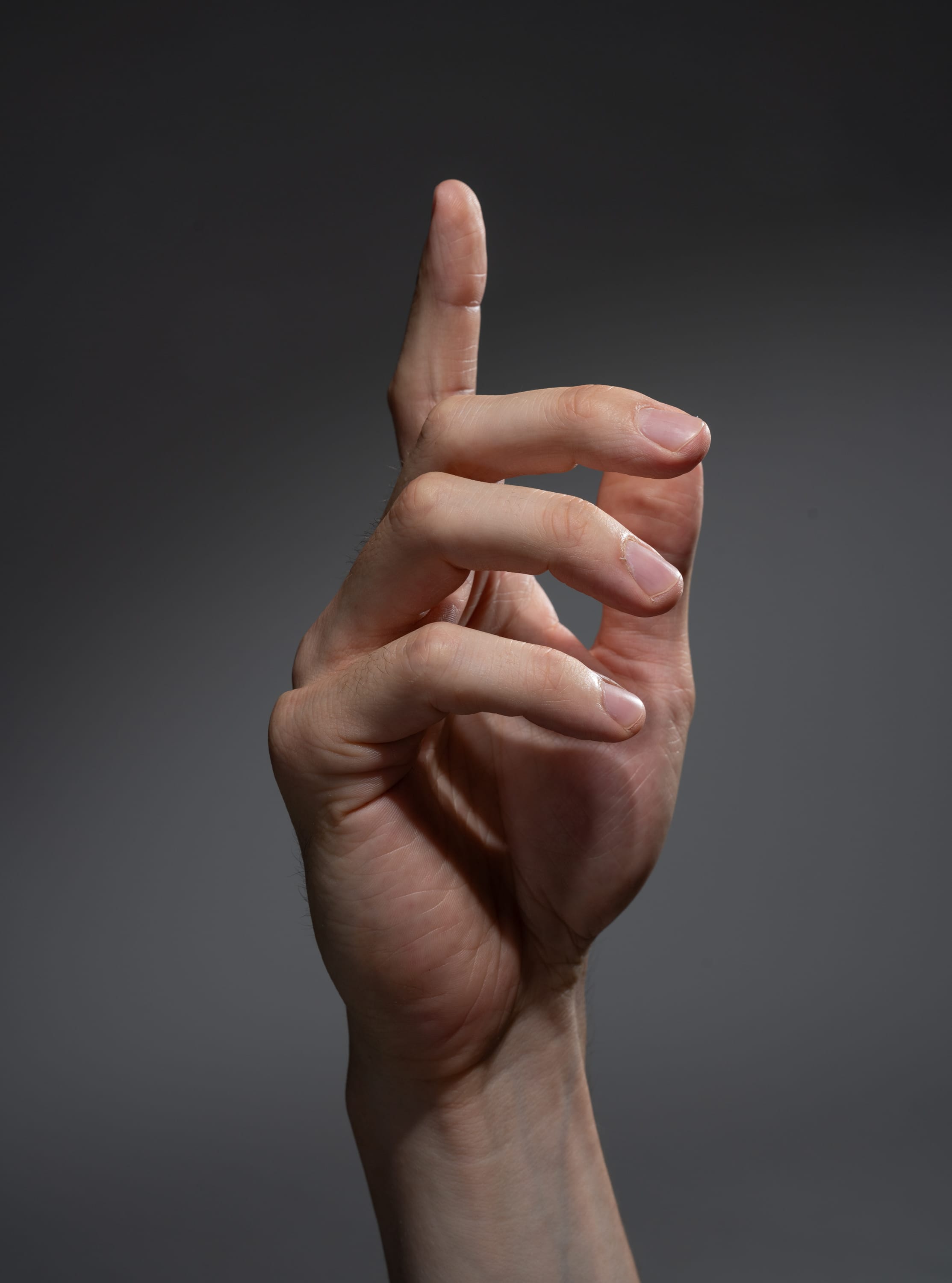 a single male hand is extended against a gray background. the hand is lit from the side, creating distinct shadows and highlights. the index finger is pointing upwards, while the other fingers are slightly curled.