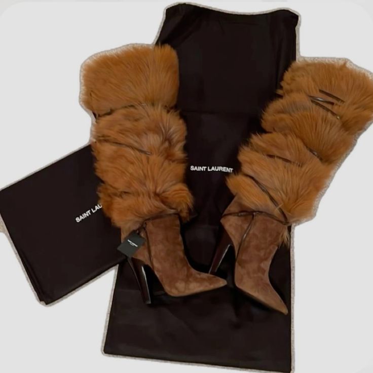 this image showcases a pair of saint laurent furlined suede boots in a rich brown hue. the boots feature a luxurious fur trim and are presented with a black dust bag. the dust bag prominently displays the brand name, "saint laurent," adding an authentic touch to the presentation. the boots, characterized by their high heels and sleek anklefitting design, exude a sense of style and elegance, suggesting a premium fashion piece for the winter or fall season.