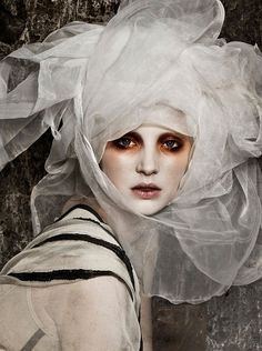 a woman with her face partially covered by a white veil, her eyes are heavily made up with dark eyeliner and orange eyeshadow. she is wearing a white and black patterned dress. the photo is taken in a studio setting with a plain background.