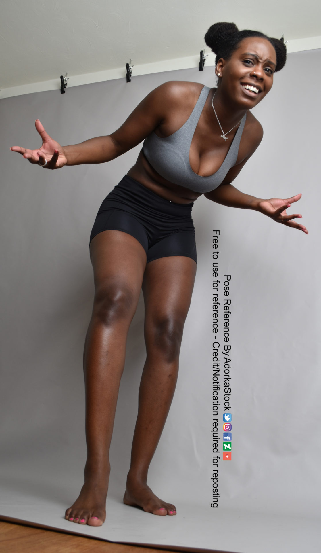 a woman in a sports bra and shorts stands with her arms outstretched, smiling, with a casual look on her face. she is facing the camera, and is standing on a white background.  her legs are apart, and her arms are slightly raised.  her expression is friendly and approachable.