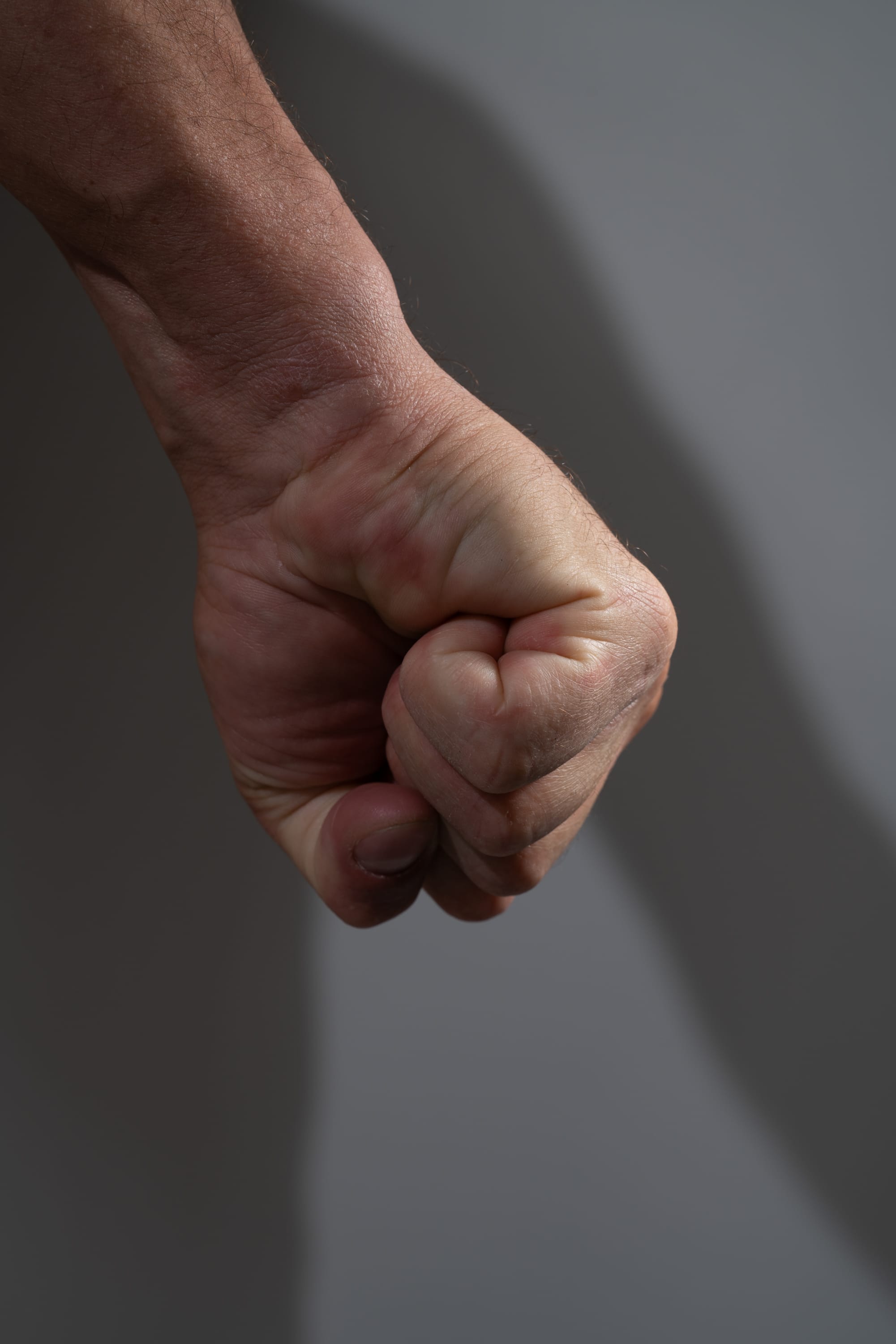 the image shows a male hand clenched in a fist, set against a gray background. the hand is illuminated by studio lighting, creating a strong contrast of light and shadow. the closeup perspective emphasizes the tension and power of the clenched fist.