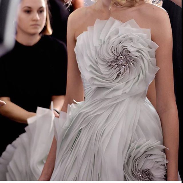 a woman wearing a stunning white gown adorned with intricate ruffles and floral designs stands in a glamorous setting. the dress, with its flowing fabric, exudes elegance and sophistication. silver accents and jewelry add a touch of sparkle to the ensemble. the woman's confident posture and radiant expression suggest a sense of confidence and beauty.