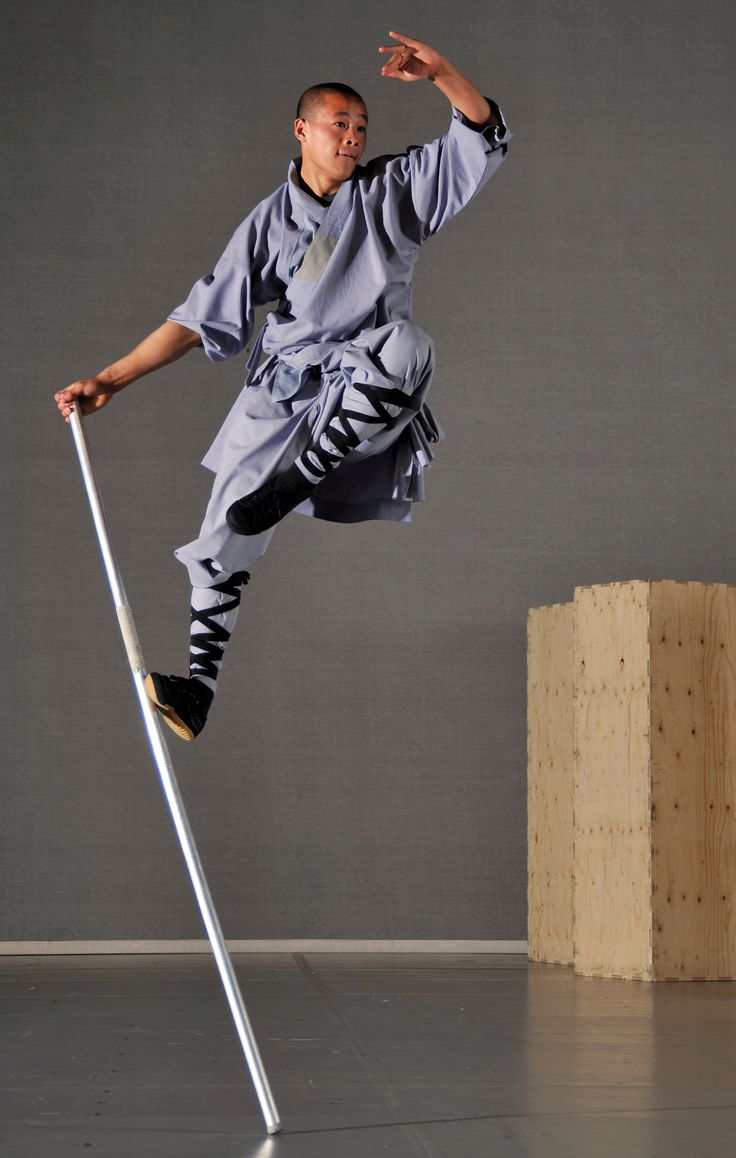 a young man, dressed in a grey shaolin monk uniform, is midair while jumping over a pole. he is midkick and looks like he is training in martial arts.  the man is against a plain grey wall and the pole is the only other object.