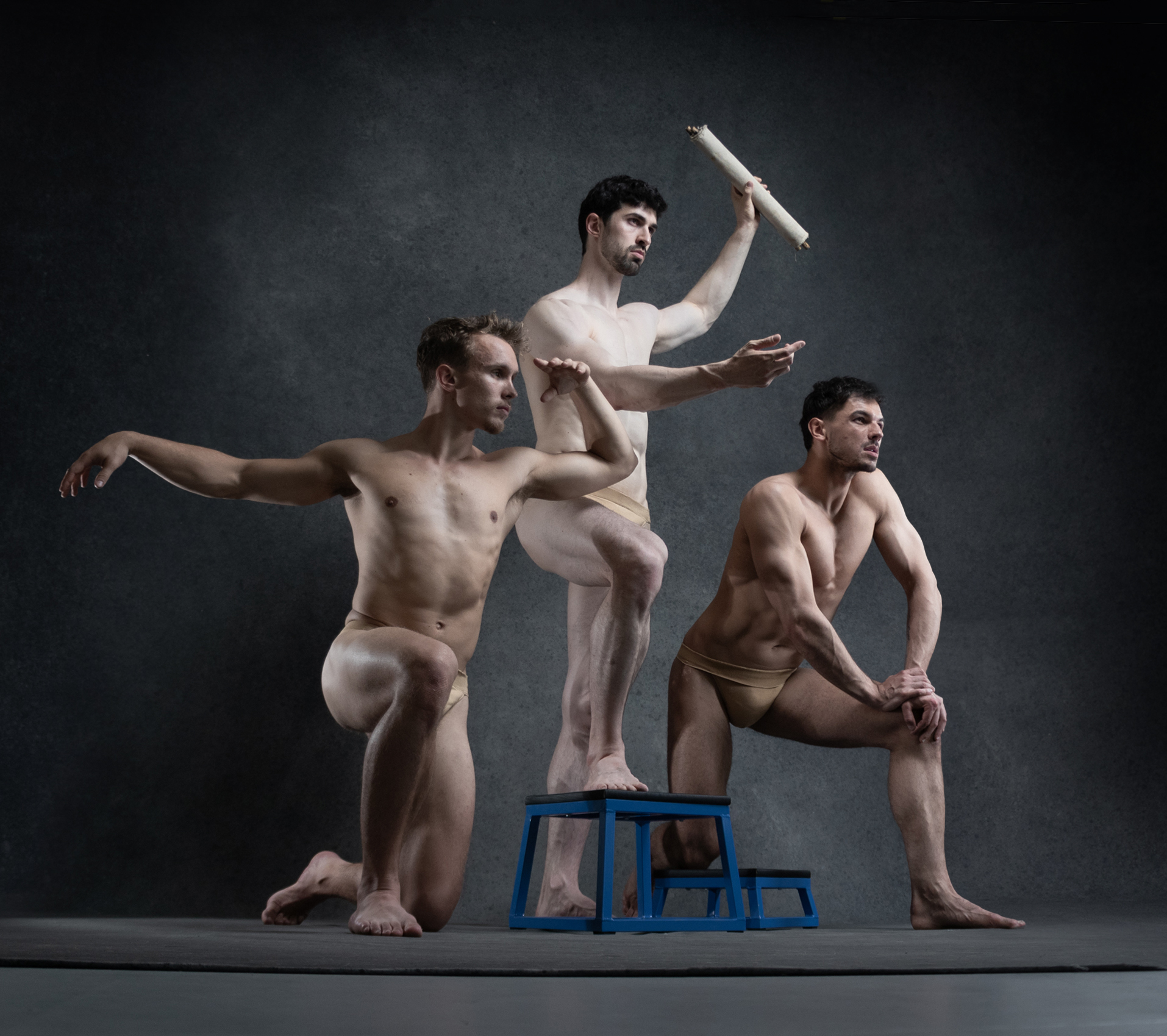 three muscular men with toned physiques are captured in dynamic dance poses against a dark, textured background. the lighting accentuates their muscles and the fluidity of their movements.  the man on the left extends his arm gracefully, while the central figure balances on a small platform, his body forming an elegant arc. the man on the right adopts a crouched position, his gaze intense and focused. the image evokes a sense of strength, athleticism, and artistic expression.