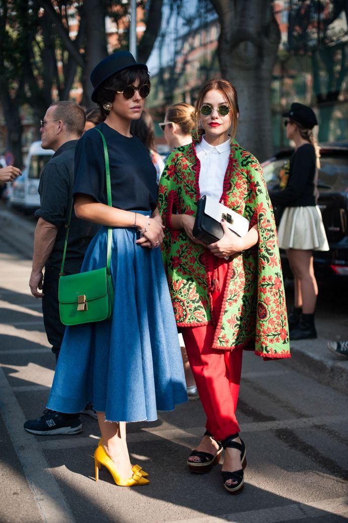 two stylish women are walking down a city street.  the woman on the left wears a black hat, sunglasses, a navy blue top, and a full denim blue skirt. she carries a bright green shoulder bag and is wearing yellow high heels. the woman on the right wears a white shirt, vibrant red pants, and a richly patterned green and red cape.  she's also wearing sunglasses and carries a white and black handbag.  both women are welldressed and display a trendy street style.  other people, some slightly blurred, are in the background, suggesting a bustling city scene. the overall atmosphere suggests a fashion event or stylish urban setting.