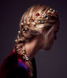 a woman with long blonde hair styled in a side braid adorned with pearl and gem hair clips.  she is wearing a dark red velvet jacket and is looking down.  the background is a dark grey.