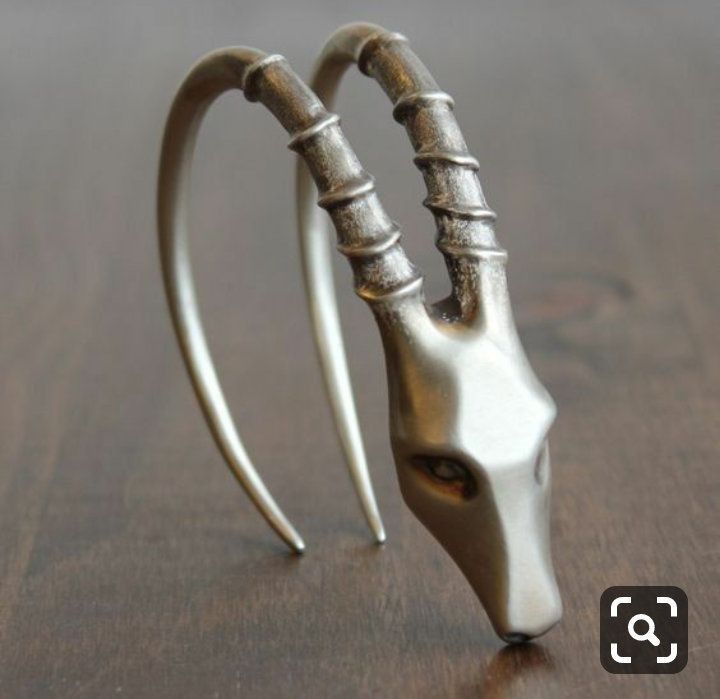 an image of a silver earring in the shape of an antelope head. the earring is made of metal and has a curved shape, with the antelope's head at one end. the antelope has long, curved horns that are detailed with ridges. the earring is sitting on a brown wooden surface.