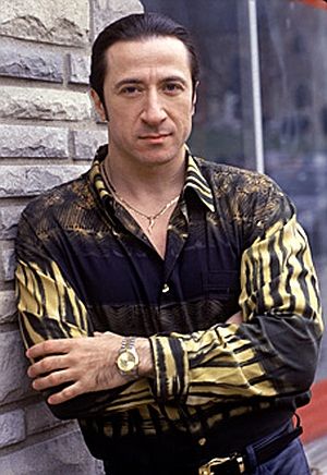 a photo of rosario fiorello is captured in a medium shot, as he leans against a brick wall outdoors. he is wearing a darkcolored, buttondown shirt with a distinctive pattern in yellow. adorning his neck is a delicate gold chain. on his wrist, he wears a gold wristwatch. his hair is neatly slicked back, adding to his polished appearance. his arms are crossed in front of him. he is wearing a belt.