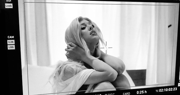 a black and white portrait of a blonde woman with her hands behind her head, looking up. the image is captured on a monitor, displaying camera settings and timecode information. a focus point is visible in the center of the frame.