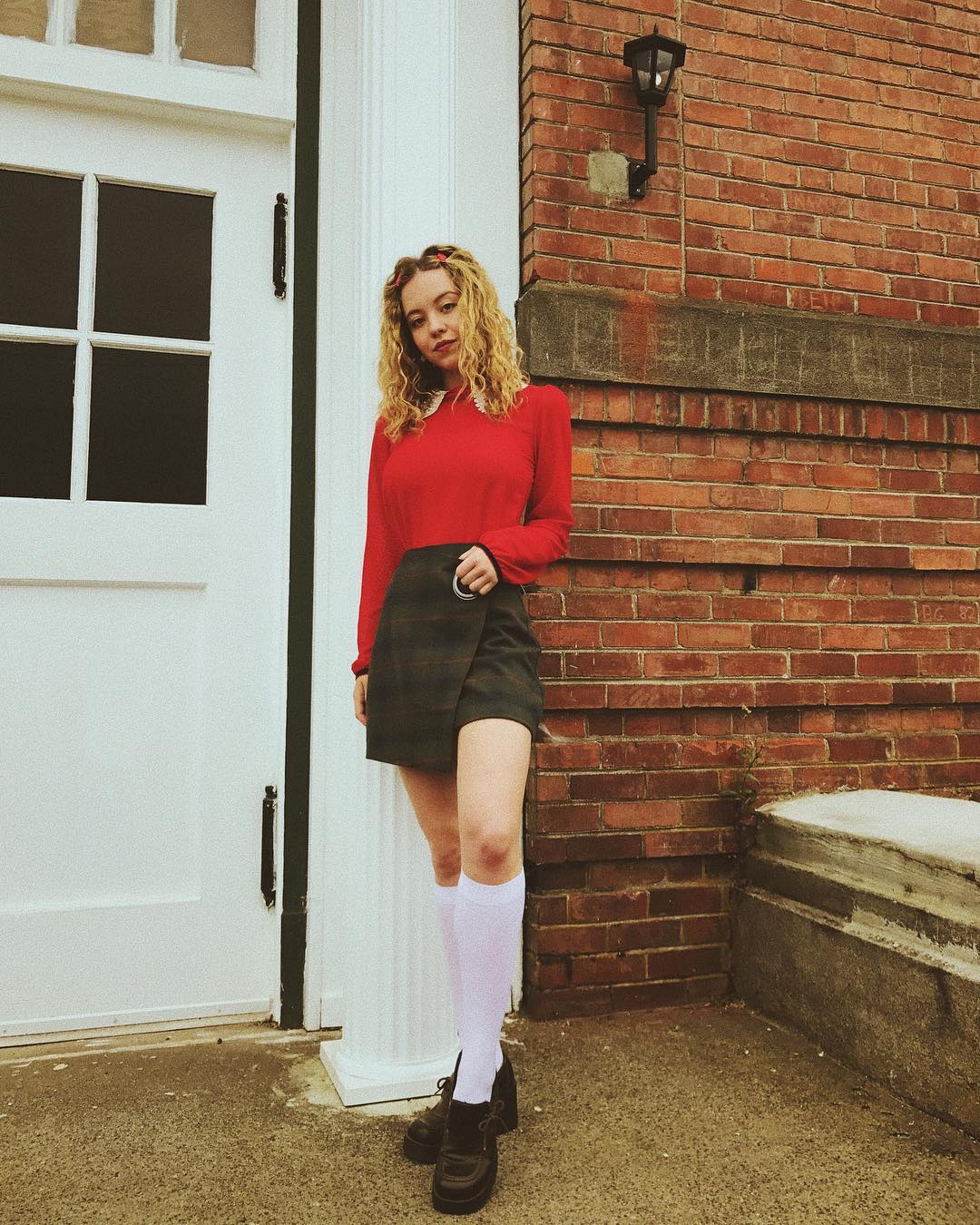 the image shows a young woman standing against a brick wall next to a white door. she's wearing a red longsleeved sweater, a dark green and brown plaid wraparound skirt, white kneehigh socks, and black chunky shoes.  her hair is curly and blonde. the overall style is reminiscent of 90s or vintage fashion. the scene is outdoors, possibly near a school or older building.
