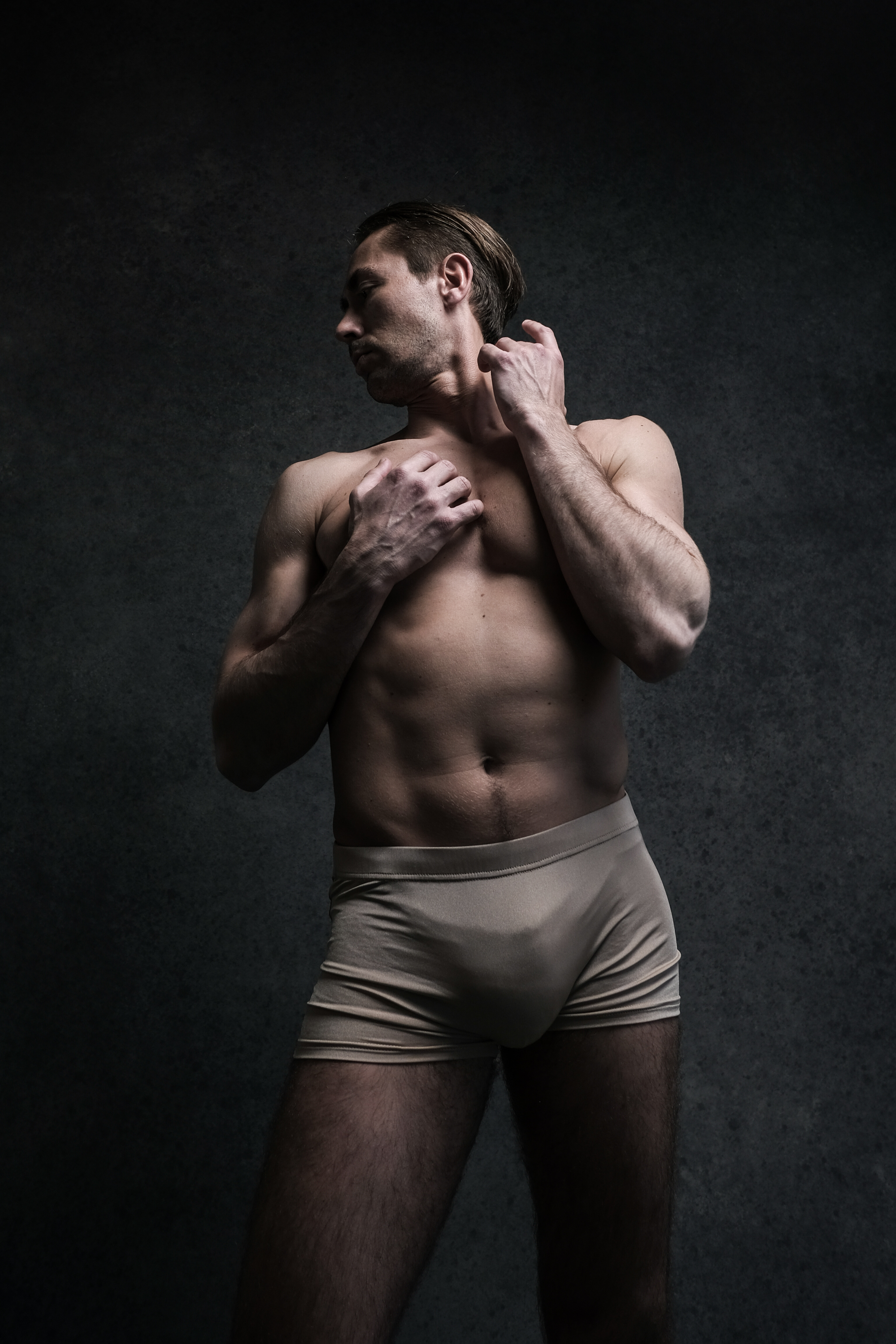 a shirtless man with a toned physique stands against a dark background. he is wearing lightcolored underwear and is lit with dramatic studio lighting. the man is striking a pose, with one hand on his chest and the other behind his head, creating a sense of confidence and contemplation.