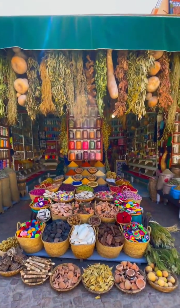 a vibrant market stall overflows with a colorful array of spices, herbs, and other goods. baskets filled with various items line the front, while shelves behind showcase spices in jars and dried goods. dried herbs and gourds hang from the awning above.