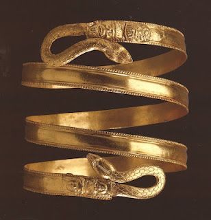 an ancient gold spiral armband or bracelet depicting a snake wrapped around the arm. the band is relatively wide and smooth, and the snake head and tail are visible at either end. there are decorative details on the head and tail. the piece appears to be photographed against a dark brown background.