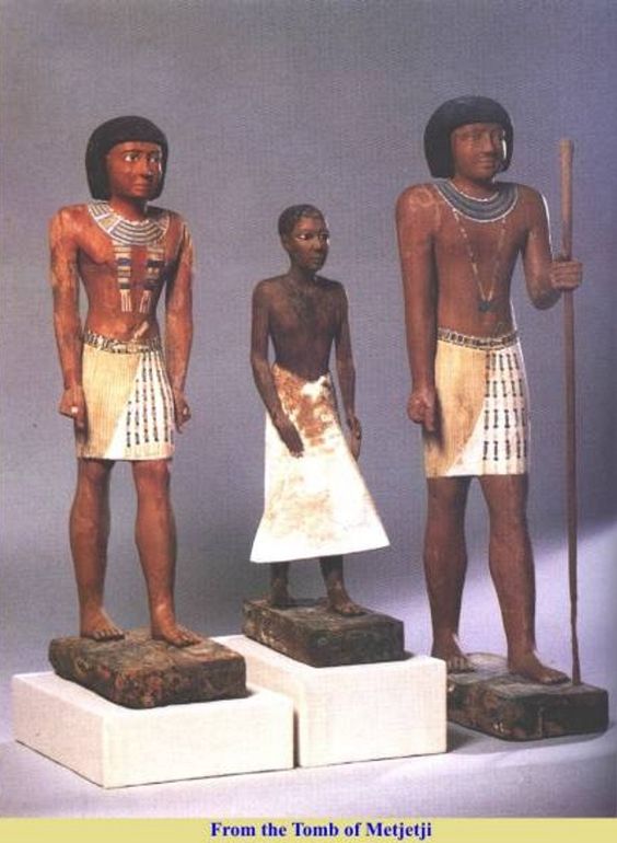 three wooden statues from the tomb of metjetji in ancient egypt. two taller figures flank a shorter, darker figure in the center. all three are adorned with necklaces and kilts. the two taller figures are similar in appearance, with reddishbrown skin, black hair, and a staff held by the figure on the right. the central figure has darker skin and is dressed in a white kilt. the statues stand on rectangular bases.