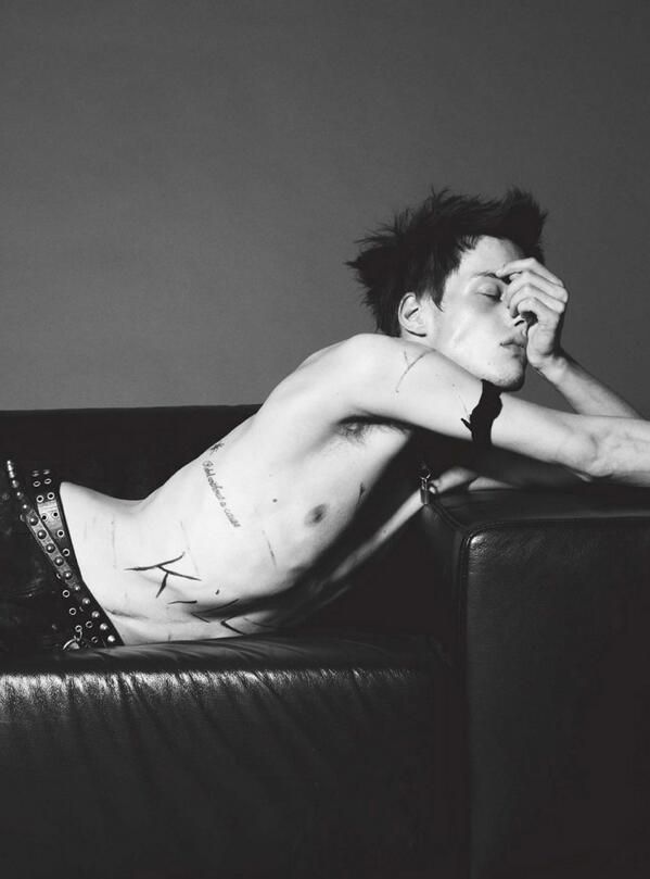 a black and white portrait of a young, handsome man. he is sitting on a leather couch and appears to be shirtless. the man has a tattoo on his side and his hair is styled in a messy, edgy way. the image has a cool, moody aesthetic.