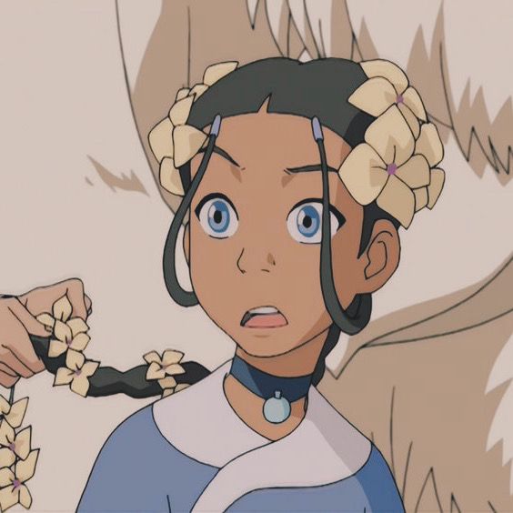 katara from avatar: the last airbender is looking up to the right with a concerned expression. she has long dark hair, brown skin, light blue eyes, and a flower braid headband. katara is wearing a blue tunic with a light blue trim and a light blue necklace with a turquoise pendant. she is standing in front of a white and brown background.