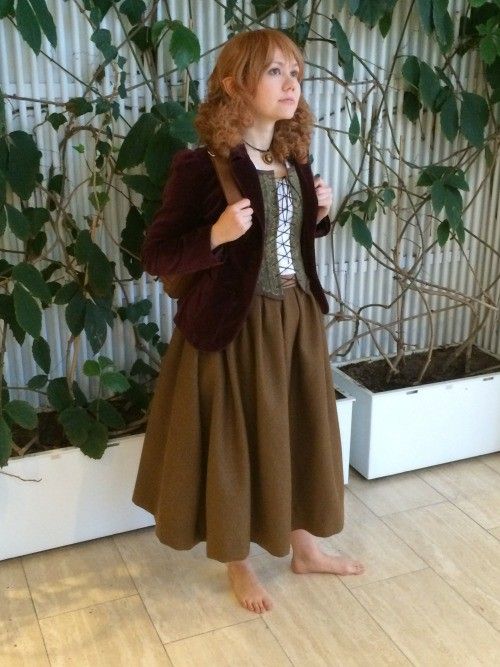 a young redhead woman is cosplaying as an elven or hobbitlike character. she is wearing a brown long skirt, a burgundy velvet jacket, a laced corsetstyle top, and a brown backpack. she is barefoot and standing in front of a wall with ivy plants. she is looking up and slightly to the side. the setting appears to be indoors. the overall aesthetic is whimsical and fantasyinspired.