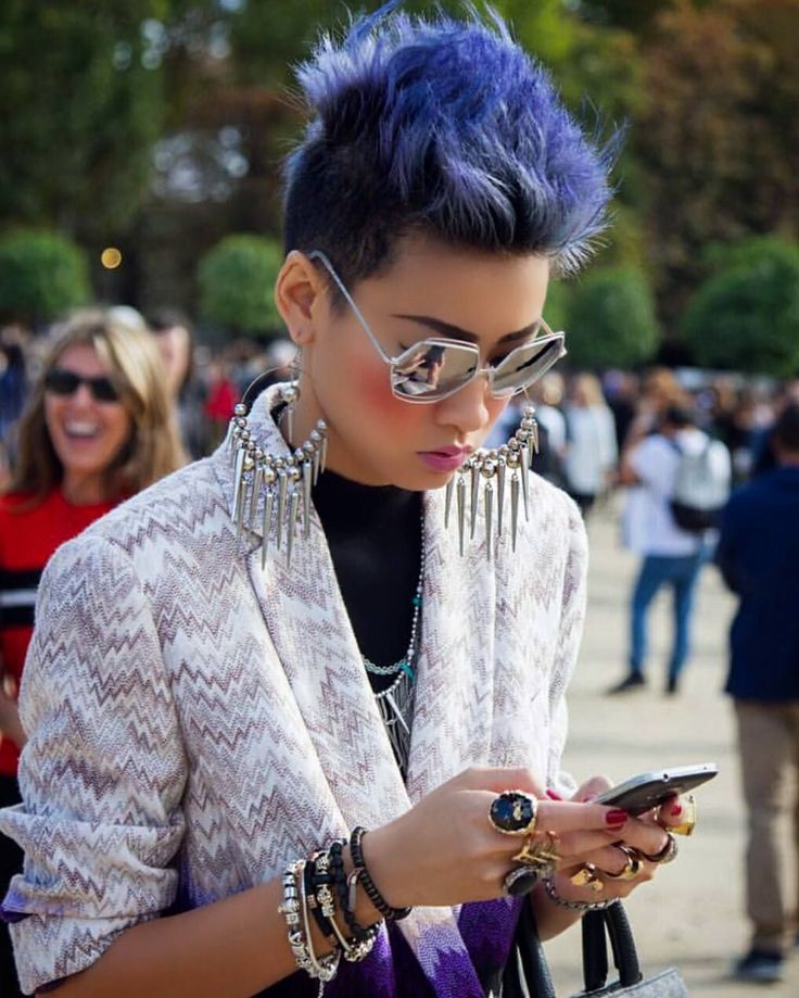 a fashionable woman with blue hair is looking at her phone. she is wearing sunglasses, large earrings, and several bracelets and rings. she is wearing a patterned blazer over a black turtleneck. a crowd of people is in the background.
