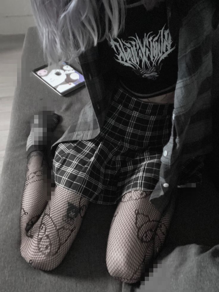 a woman is sitting on a bed. she has gray hair and is wearing a black crop top with a white design on it, a plaid skirt, a flannel shirt, and fishnet stockings. a phone is resting on the bed next to her. parts of the image have been pixelated to obscure some details.