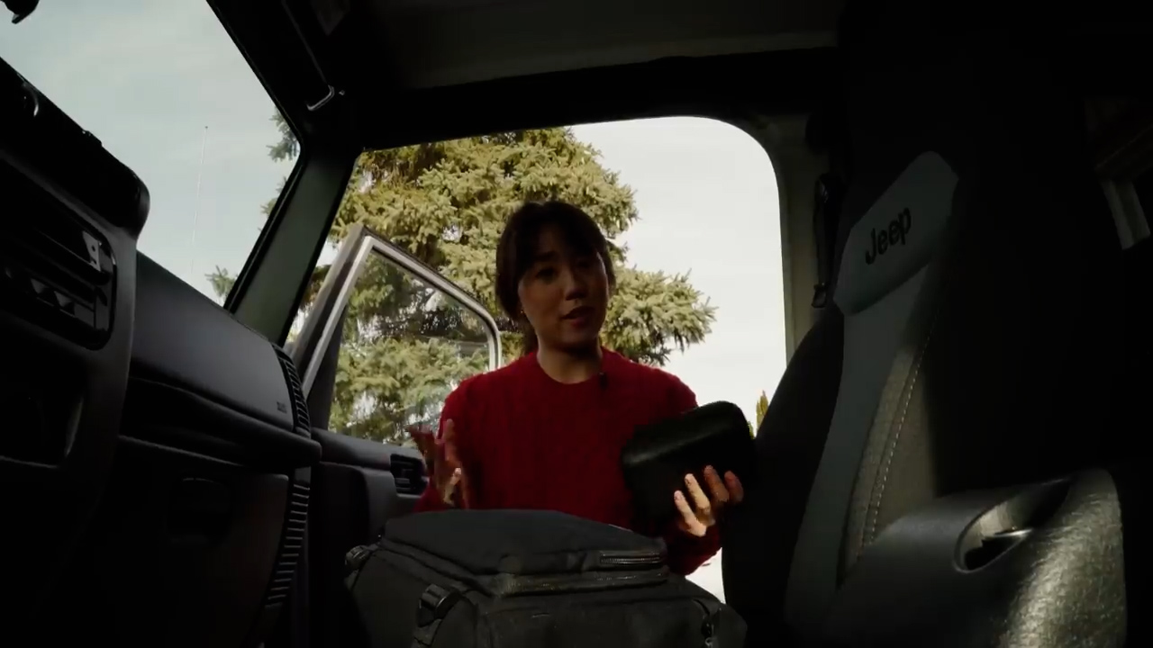a woman stands next to the open door of a jeep, partially inside the vehicle. she is holding a small black bag in her left hand and gesturing with her right hand as if talking to someone. a black backpack sits on the passenger seat beside her. the car appears to be parked outdoors, with trees visible through the windshield and open door. the lighting is bright and natural, suggesting a sunny day. the perspective is from inside the car, looking out.