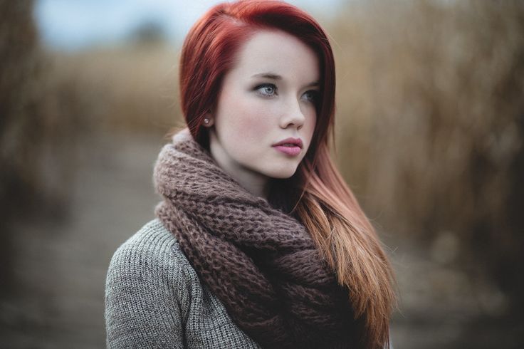 a young woman with long red hair and a brown scarf is photographed outdoors in a field. she is wearing a gray sweater and looking off to the side. the lighting is soft and natural, giving the image a serene feel.  the colors are muted and earthy, with browns, grays, and tans dominating the scene.