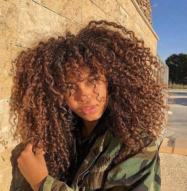 a portrait of a woman with voluminous, brown, curly hair and bangs, wearing a camouflage jacket. she stands outdoors near a building, with natural light casting shadows and highlighting her complexion. the image has warm tones and focuses on her beauty and style.