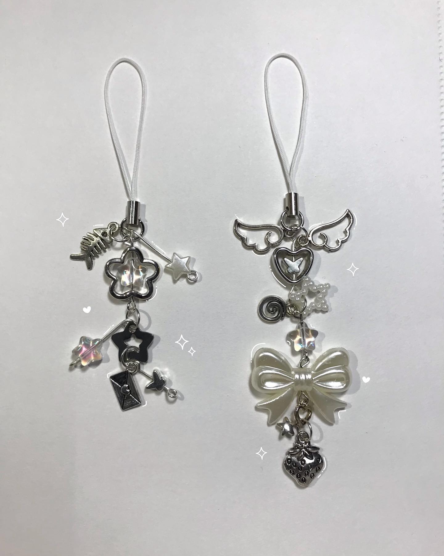 the image shows two phone charms on a white background. the charm on the left has a white loop at the top and dangling from it are a silver fish skeleton, flower, star, heart, envelope, and other small starshaped pieces. the charm on the right has a white loop at the top and dangling from it are silver wings, a heart, a swirl, a pearl bead, a bow, a star, and a strawberry.