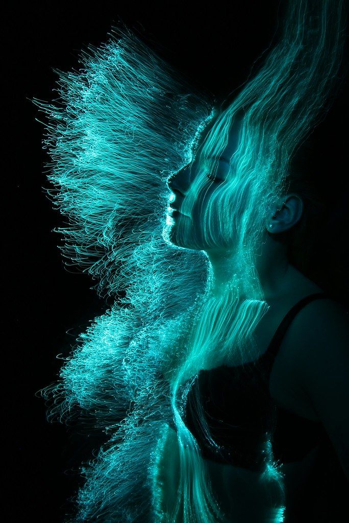 the image is a striking light painting portrait of a woman.  her profile is visible, but her form is largely obscured and redefined by streaks of vibrant turquoise light, which appear to be created using fiber optics or a similar technique. the light creates a flowing, almost ethereal effect, making her look otherworldly. the background is completely black, allowing the luminous teal strands to stand out dramatically. the overall impression is one of movement, energy, and mystical beauty.