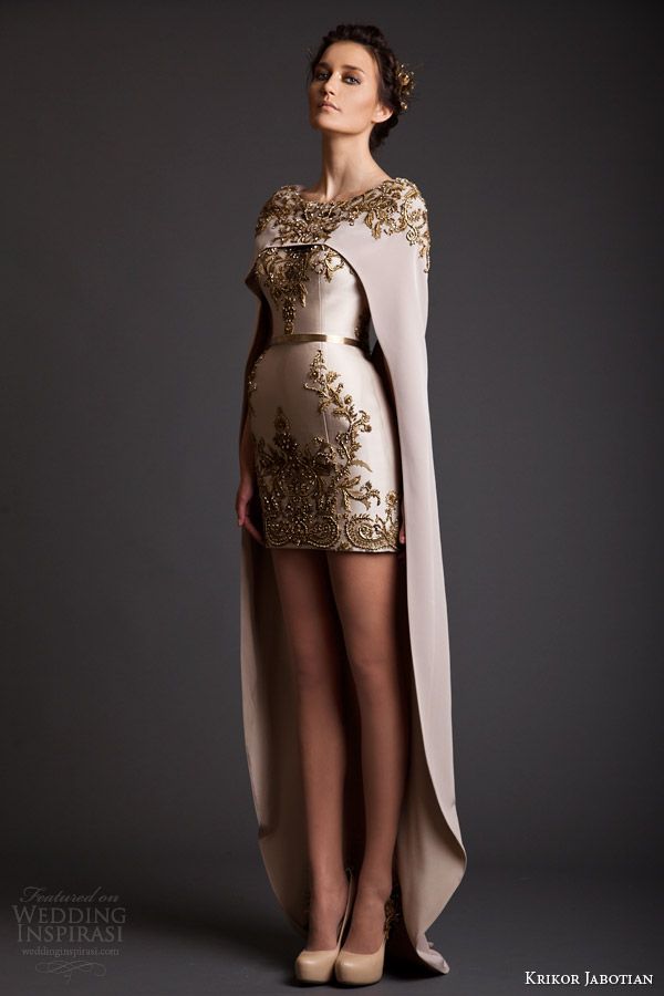 a full shot of a model wearing a beige, short, embellished wedding dress with a long cape. the dress is intricately embroidered with gold thread and features a fitted bodice and a slightly aline skirt. the model is wearing nude heels and has her hair styled up. the background is a dark grey studio backdrop. the image is professionally photographed, highlighting the details of the dress and the model's pose.  a watermark indicates that the image is featured on wedding inspirasi. the designer's name, krikor jabotian, is also visible in the bottom right corner.