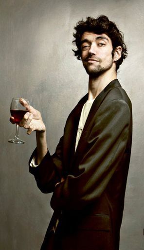 a man in a black jacket is holding a glass of red wine and looking off to the side. he is standing against a plain gray background. he has a serious expression on his face. he is wearing a black suit and a white shirt. the photo is lit from the right side.  the shadow from his jacket and his hand are visible on the wall.