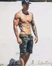 the image features a muscular man with tattoos standing in front of a white wall. he is wearing camouflage shorts and a black baseball cap. the lighting is sunny and natural, creating a bright and airy feel.