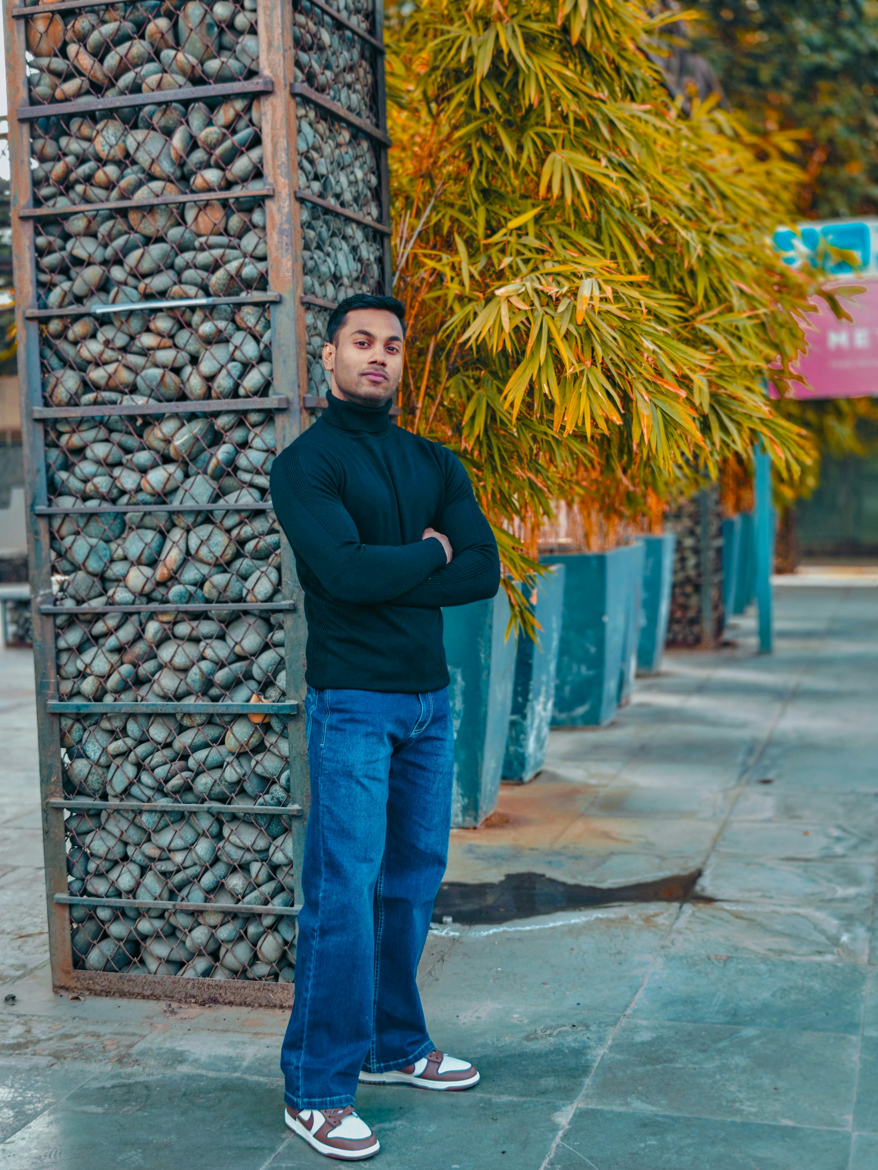 a man is standing outdoors with his arms crossed. he is wearing a black turtleneck, blue jeans, and sneakers. he is standing next to a structure filled with rocks and in front of a row of bamboo trees.