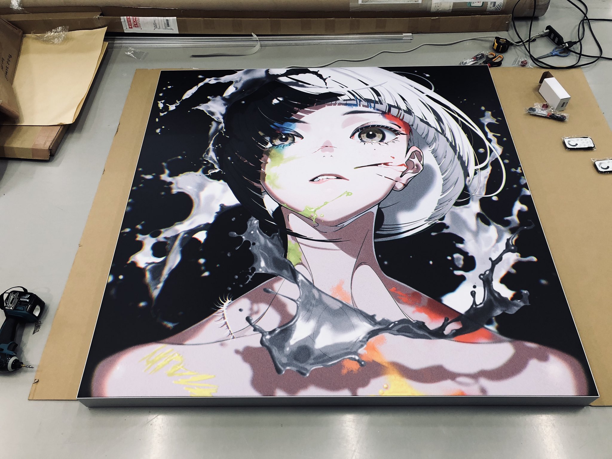 a large, almost lifesized print of an animestyle illustration lies on the floor.  the artwork features a girl with black and white hair, splashed with colorful paint, against a dark background. it appears to be freshly printed and ready for framing or mounting, as suggested by the packaging materials and tools scattered around.