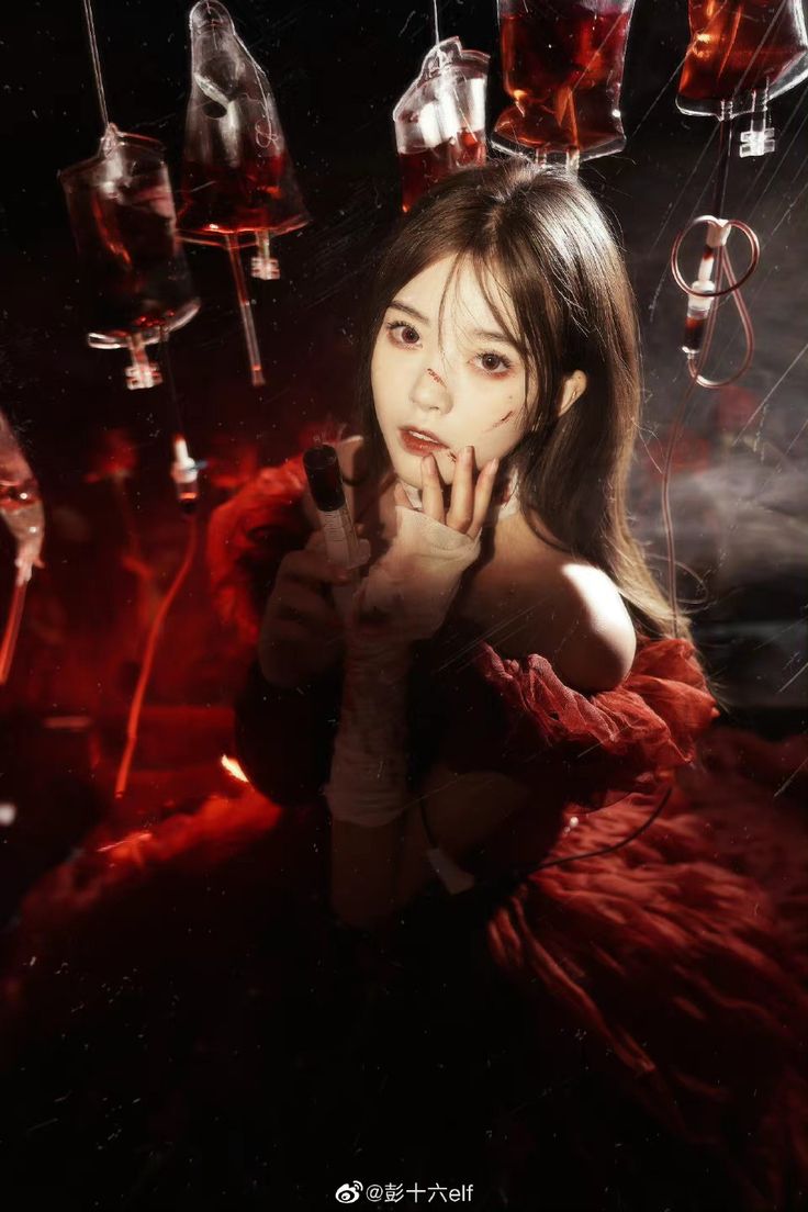 a young woman in a red dress is posed in a dark, atmospheric setting.  she has bloody makeup on her face and arms and appears to be in a bloody, spooky environment with hanging blood bags above her. she is holding something in her hands, perhaps a prop related to the theme.  the image has a gothic, horror, and somewhat unsettling tone. the lighting is dramatic, with shadows and highlights that create a mood of mystery and suspense.